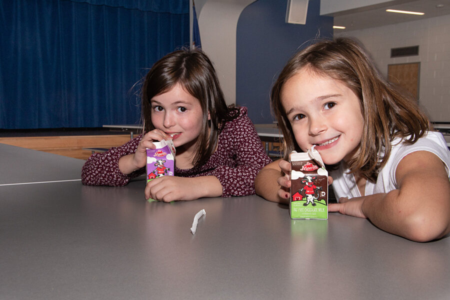 Two girls drink from half pint paper milk cartons while smiling.